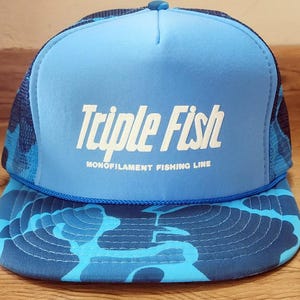 Könnte beinhalten: Eine hellblaue Trucker-Cap mit dunkelblauem Tarnmuster auf dem Netzrücken und dem flachen Schirm. Das Vorderteil zeigt den weißen Schriftzug Triple Fish und den kleineren weißen Text MONOFILAMENT FISHING LINE mit einer blauen Kordel.
