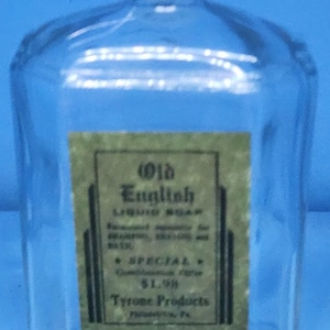 May include: A clear glass bottle with a yellow cap, labeled "Old English Liquid Soap." The label is green with black text. The bottle has a unique shape and is set against a blue background.