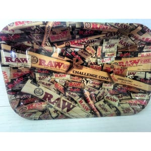 May include: A rectangular rolling tray with a colorful collage of RAW rolling paper packaging. The tray features various sizes and styles of RAW papers, with text like "CLASSIC" and "CHALLENGE CONE" visible. The tray is on a white surface.