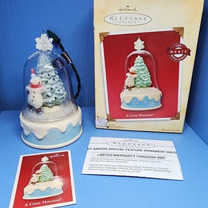 May include: A Hallmark Keepsake ornament featuring a snowman and a Christmas tree inside a glass dome. The base is light blue with snowflake details. The box and card read "A Cool Holiday!"
