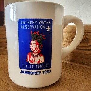 May include: White ceramic mug with a blue rectangular design. The text on the mug reads "ANTHONY WAYNE RESERVATION" and "LITTLE TURTLE JAMBOREE 1980." The mug features a Native American chief design and has a curved handle.