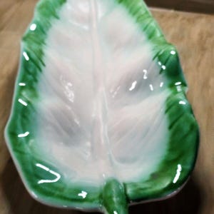 Op de afbeelding: Een keramieken schaal in de vorm van een blad met een glanzende afwerking. Het midden van de schaal is wit en gaat over in een levendige groene kleur langs de geschulpte randen. Een verhoogde centrale nerf loopt over het blad.