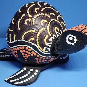 Könnte beinhalten: Handbemalte Holzschildkrötenfigur. Der Panzer ist schwarz mit gelben, weißen und kastanienbraunen Mustern. Kopf und Flossen sind schwarz mit orangefarbenen und weißen Akzenten. Ein dekoratives Objekt.