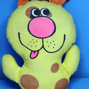 Pode incluir: Um brinquedo de pelúcia em forma de cachorro em tons de amarelo, marrom e rosa. O cachorro tem grandes manchas marrons, orelhas laranja e um nariz rosa. O brinquedo tem um rosto sorridente com detalhes pretos e uma língua rosa.
