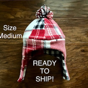 May include: A medium-sized, plaid fleece hat with ear flaps and a pom-pom. The hat features red, white, and black plaid patterns. The text "READY TO SHIP!" is displayed on the hat.