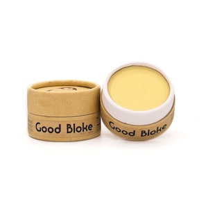 May include: Two small, round containers of balm, one open and one closed. The containers are made of brown cardboard with a white label that reads "Good Bloke". The balm is a pale yellow color.
