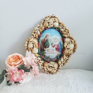 May include: A framed image of the Holy Family, Mary, Joseph, and baby Jesus, surrounded by a decorative frame made of seashells and beads.