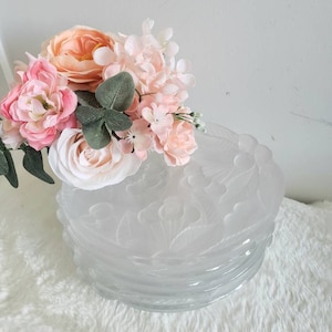 Puede incluir: Un conjunto de cuatro platos de vidrio transparente con un diseño floral en relieve. Los platos están decorados con un delicado patrón floral y tienen un borde festoneado. Los platos están cubiertos con un ramo de flores artificiales de color rosa y melocotón.