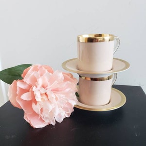 May include: Two pink teacups with gold trim and matching saucers. The cups and saucers are stacked on top of each other. A pink flower is in the foreground.
