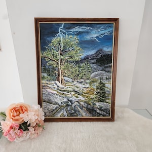 May include: A framed oil painting depicting a stormy mountain landscape with a single tree in the foreground. The sky is dark blue with streaks of lightning. The mountains are rocky and covered in snow. The tree is green and has a thick trunk. The painting is framed in a brown wooden frame.