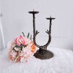 May include: A black metal candle holder with a tree branch design. The holder has two candle cups and a round base. The holder is sitting on a white fluffy surface.