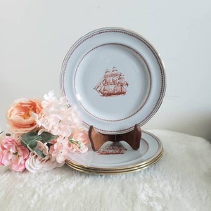 May include: Set of four white ceramic dinner plates with a gold rim and a brown ship illustration in the center. The plates are stacked on a wooden stand.