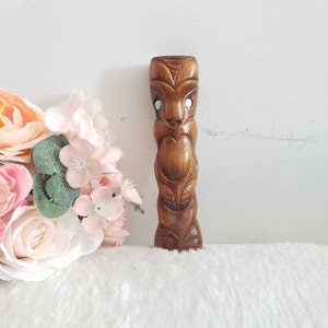 Peut inclure: Une petite sculpture en bois d'une figure tiki avec des yeux bleus. La figure est sculptée dans un style traditionnel et a une finition brune.
