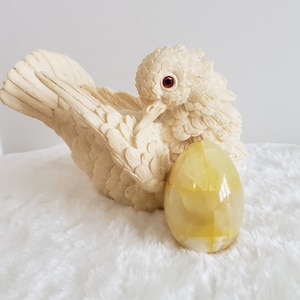 Peut inclure: Une figurine d'oiseau crème aux yeux rouges, à côté d'un œuf en pierre jaune et blanc. L'oiseau est détaillé avec des plumes texturées. L'œuf a une surface polie et repose sur une surface blanche et duveteuse.