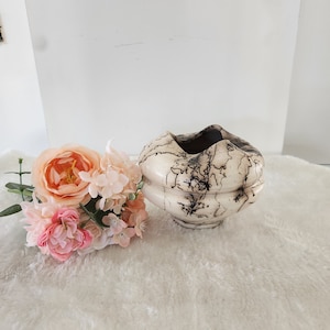May include: A white ceramic vase with a brown crackle glaze. The vase has a wide, round opening and a narrow base. It is sitting on a white fluffy surface with a bouquet of pink flowers.