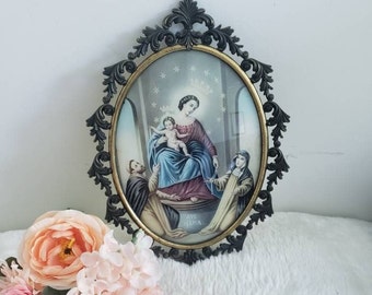 May include: An oval-shaped framed image of the Virgin Mary holding baby Jesus. The image is surrounded by a gold-coloured frame with a black ornate border. The image is set against a white background. The text "AVE MARIA" is visible at the bottom of the image.