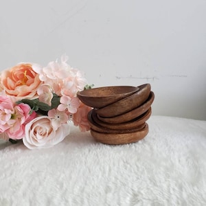 May include: A stack of eight small, round wooden bowls. The bowls are a rich brown color and are stacked on top of each other. The bowls are sitting on a white fluffy surface.