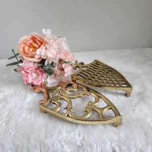May include: Two gold-toned metal trivets in the shape of a shield. The trivets have an intricate design and are perfect for protecting surfaces from hot dishes.