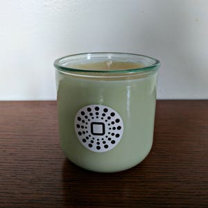 Pode incluir: Um frasco de vela de vidro verde com um rótulo branco que apresenta um círculo preto com um quadrado no centro e um padrão de pontos ao redor do quadrado.