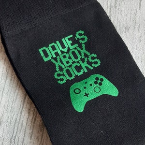 Könnte beinhalten: Schwarze Socken mit grünem Text, der "Dave's Xbox Socks" lautet, und einer grünen Grafik einer Videospielkonsole.