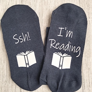 May include: A pair of navy blue socks with white text and book graphics. One sock says "Ssh!" and the other says "I'm Reading". The socks are made of a soft, comfortable material.