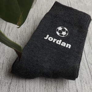 Könnte beinhalten: Anthrazitfarbene Socken mit einem weißen Fußball-Grafik und dem Namen "Jordan" darunter. Die Socken sind gefaltet und scheinen aus einem weichen Material zu sein. Ein grünes Pflanzenblatt ist im Hintergrund zu sehen.