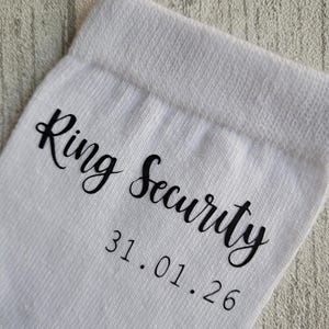 May include: White sock with the words "Ring Security" in a black script font. Below the text is the date "31.01.26" in a smaller, black font. The sock is made of a textured, knit fabric.