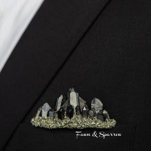 Op de afbeelding: Zwarte blazer met zichtbaar wit overhemd. De zak toont een broche met een bergontwerp van kristallen en een basis van kleine, goudkleurige stenen. De tekst "Fawn & Sparrow" is aanwezig. Een stijlvol accessoire.