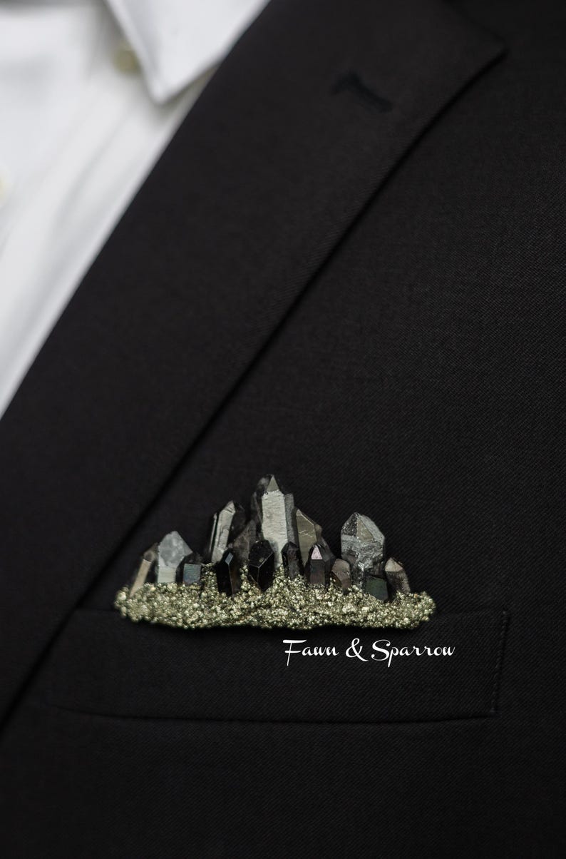 Op de afbeelding: Zwarte blazer met zichtbaar wit overhemd. De zak toont een broche met een bergontwerp van kristallen en een basis van kleine, goudkleurige stenen. De tekst "Fawn & Sparrow" is aanwezig. Een stijlvol accessoire.