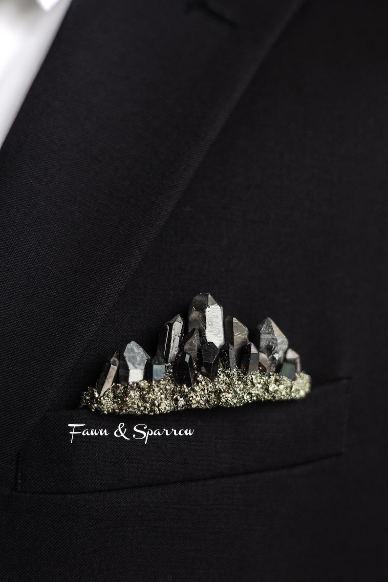 Op de afbeelding: Een zwarte kristallen broche, die lijkt op een bergketen, zit in een zak van een pak. De broche heeft donkere, facetgeslepen kristallen op een bed van metallic, goudkleurige stenen. De tekst "Fawn & Sparrow" staat eronder.