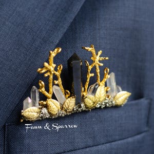 May include: A gold-toned pocket square accessory featuring a central dark crystal point flanked by clear quartz points, gold-toned branch structures, and metallic leaf accents. The text "Faun & Sparrow" is printed in white on the navy blue fabric.