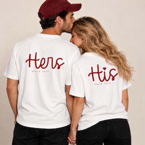 Könnte beinhalten: Zwei weiße T-Shirts mit rotem Schreibschrift-Text auf der Rückseite. Ein Shirt zeigt das Wort Hers mit dem Text SINCE 2010 darunter. Das andere Shirt zeigt das Wort His mit einem kleinen Herzsymbol und dem Text SINCE 2010 darunter.