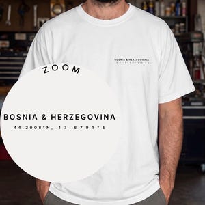 以下が含まれることがあります： 胸元に黒い文字で「BOSNIA & HERZEGOVINA」と座標「44.2008°N, 17.6791°E」がプリントされた白い半袖Tシャツ。円形のズームオーバーレイには、同じテキストと座標がより大きなフォントサイズで表示されています。