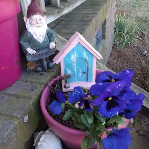 May include: A pink and blue miniature fairy house with a door and a welcome sign sits in a pink pot with purple pansies. A garden gnome with a red hat stands next to the pot.