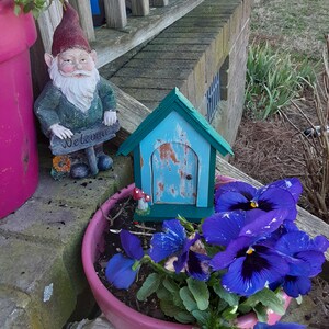 May include: A pink flower pot with purple pansies and green leaves. A small blue wooden house with a weathered door sits in the pot. A garden gnome with a red hat and a white beard stands on a wooden sign that says "Welcome".