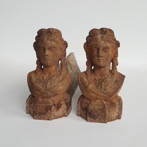 Puede incluir: Dos bustos de mujeres de estilo antiguo, con rasgos faciales detallados y cabello trenzado. Los bustos son de color marrón desgastado y descansan sobre bases rectangulares. Fondo blanco.