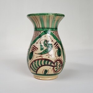 May include: A ceramic vase with a cream-colored base and green and brown painted designs. The vase features a flared rim with vertical green stripes. The body of the vase has a bird and floral motif. The vase is approximately 6 inches tall.