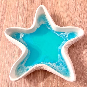 Può includere: Un piatto bianco a forma di stella marina con un bordo strutturato. L'interno è riempito di una vibrante resina turchese, che imita l'oceano. Il piatto è un oggetto decorativo.