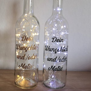 Könnte beinhalten: Zwei klare Glasflaschen mit Korkverschlüssen und weißen Lichterketten im Inneren. Jede Flasche hat einen goldenen Aufkleber mit der Aufschrift "Dein Wunschtext und/oder Motiv".