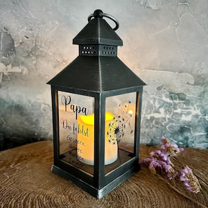 Op de afbeelding: Zwarte metalen lantaarn met glazen panelen, met een brandende kaars en de Duitse tekst "Papa, Du fehlst" (Papa, ik mis je). Een paardenbloemafbeelding en paarse bloemen zijn ook zichtbaar.