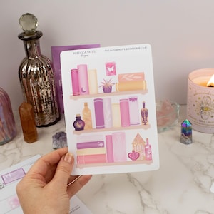 May include: A sticker sheet featuring a whimsical illustration of a bookshelf with books, bottles, and decorative elements in shades of pink, purple, and gold. The text "The Alchemist's Bookcase" is visible. Other items include a candle and crystals.