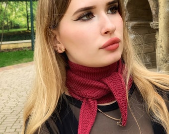May include: A deep burgundy triangular knit scarf featuring a textured garter stitch pattern. The scarf is tied in a simple knot at the front, with two pointed ends hanging down. The knit fabric is uniform in color and shows a consistent, dense stitch.