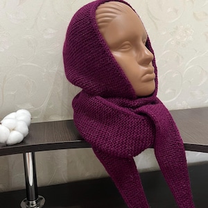 May include: A deep magenta knitted hood with an integrated scarf, featuring a textured garter stitch pattern. The garment covers the head and wraps around the neck, with two long, pointed ends hanging down in the front, crafted from yarn.