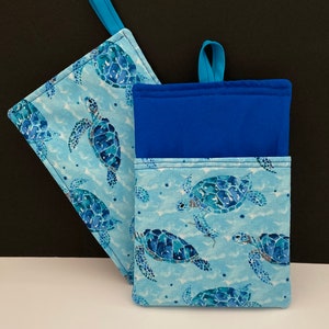 May include: Two blue and white pot holders with a sea turtle pattern. The pot holders have blue fabric loops for hanging.