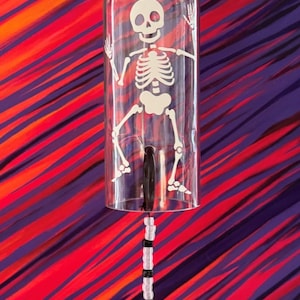 May include: A clear glass bottle wind chime featuring a white skeleton graphic and the text Glow Skeleton. The bottle is topped with a cork and includes a dangling string of alternating black and white beads ending in a small plastic skull.