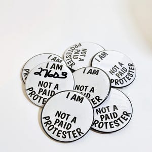 May include: A close-up of several white round buttons with black text that reads "I am not a paid protester".