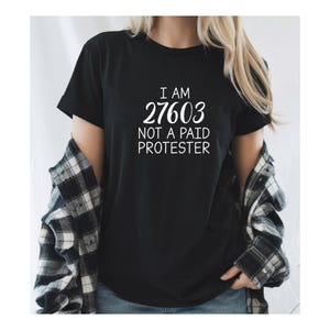 May include: Black t-shirt with white text that reads "I am 27603 not a paid protester".