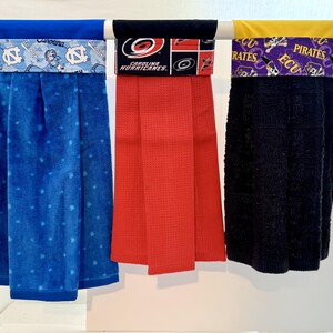 May include: Three hanging kitchen towels. The first is blue with white polka dots and a blue and white patterned top. The second is red with a textured weave and a black and red patterned top. The third is black with a yellow and purple patterned top.