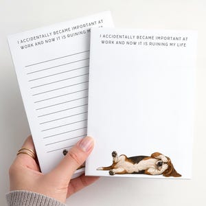 May include: Two white notepads with the text "I ACCIDENTALLY BECAME IMPORTANT AT WORK AND NOW IT IS RUINING MY LIFE". One notepad has lined pages, the other has a dog illustration. The dog is brown, black, and white.