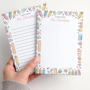 Può includere: Due blocchi per appunti con un bordo colorato di matite, gomme e righelli. Il testo "A note from Ms. Christina" è stampato in alto. Un blocco per appunti ha carta a righe, l'altro è vuoto.